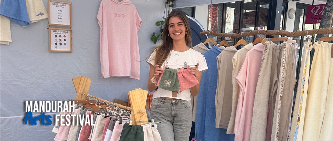 A woman stands in a market stall surrounded by hand-crafted clothing, holding a pair of cihldren's shorts and smiling
